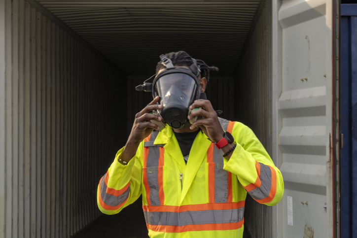 A man equipped with a powered air purifying respirator is positioned within a shipping container, showcasing safety protocols in action.