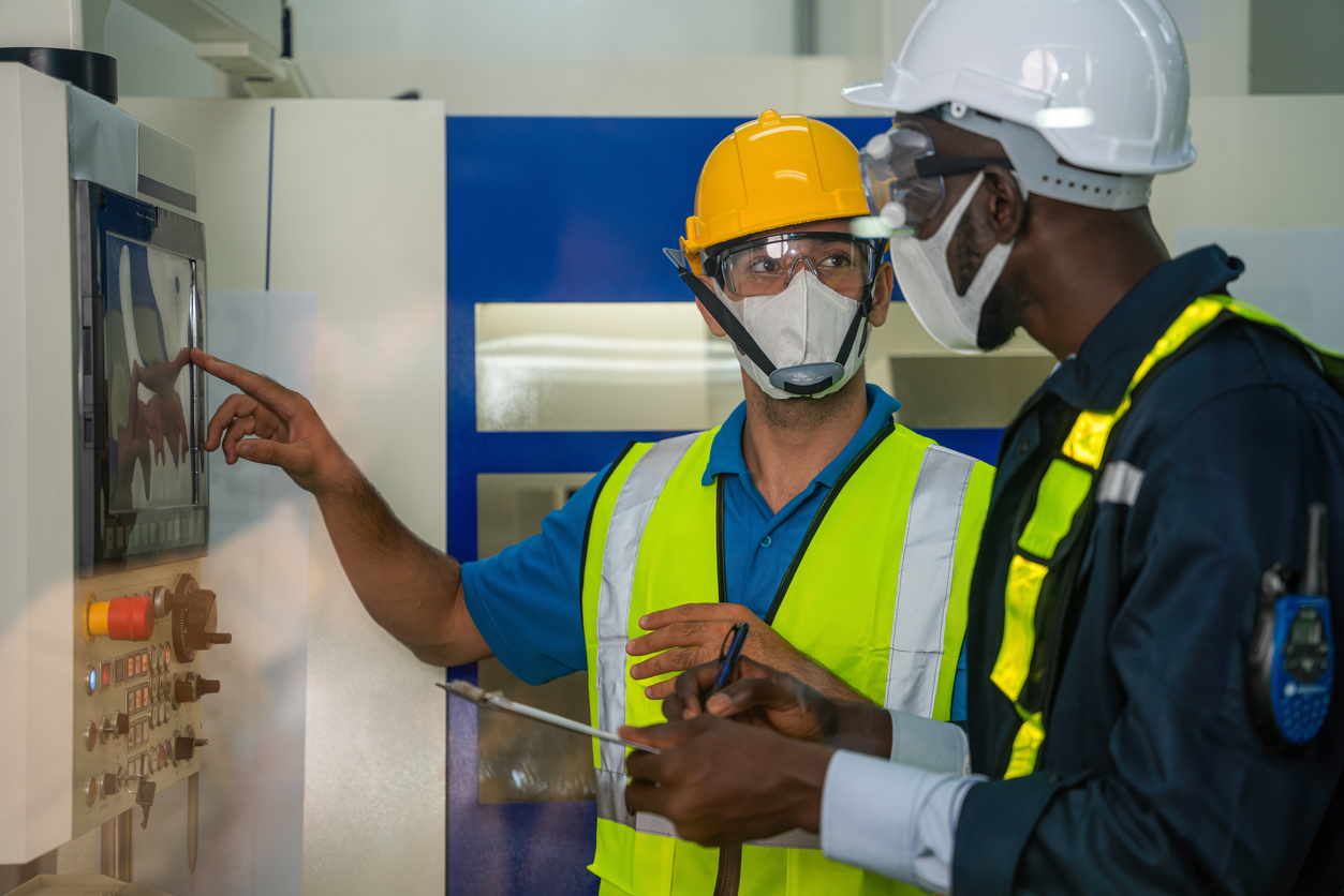 Two men in safety gear and dust masks working on a machine in an industrial setting.