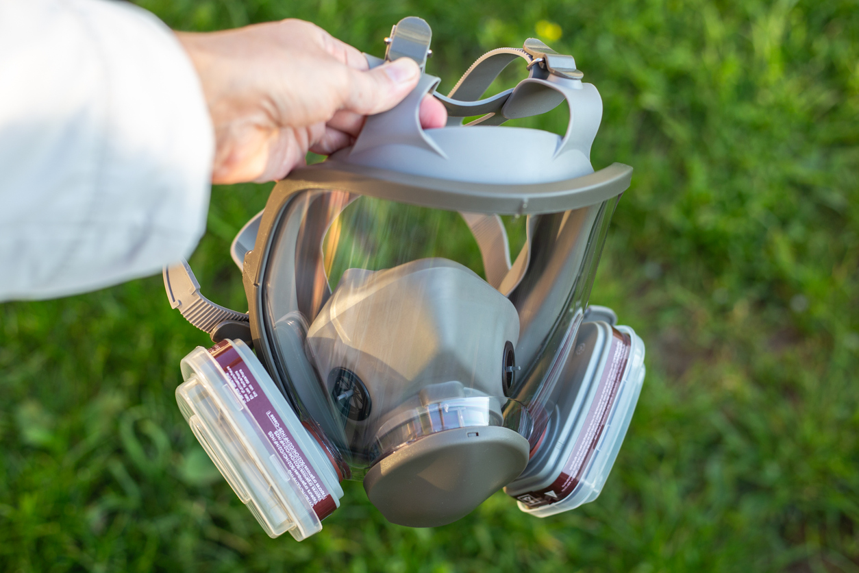 In a green grassy field, a person with a gas mask holds a reusable mask, emphasizing safety and environmental awareness.
